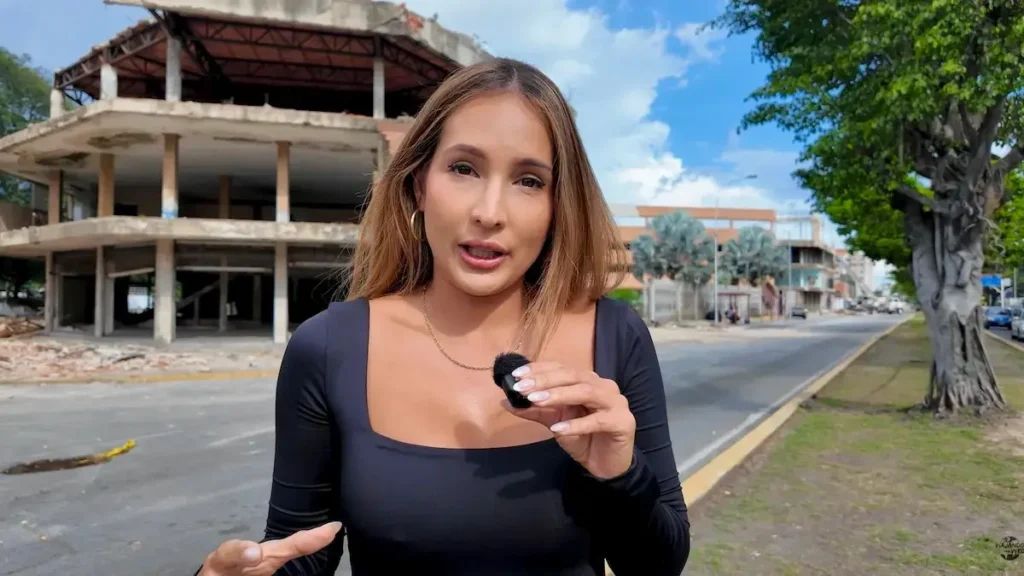 Primer plano de Vero, embajadora de Red Rumbo, reportando desde la calle con un micrófono, con un edificio de varios pisos en ruinas y colapsado por el terremoto a sus espaldas en Venezuela.