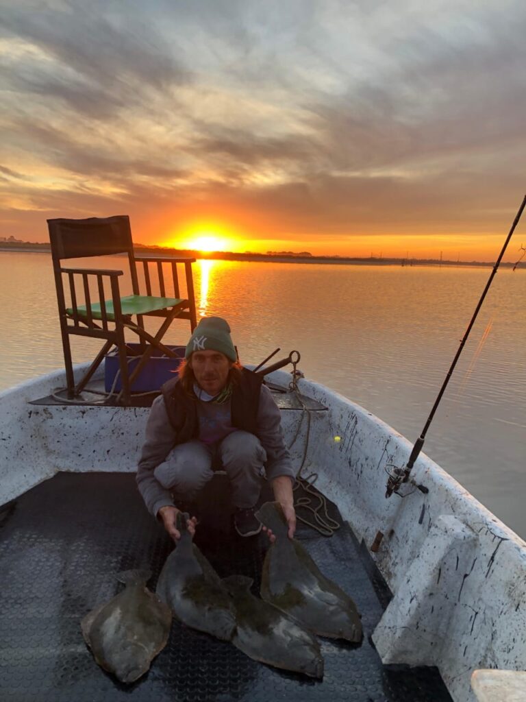"Atardecer dorado sobre la albufera de Mar Chiquita visto desde la proa de un bote, con reflejos anaranjados en el agua calma y el horizonte despejado."