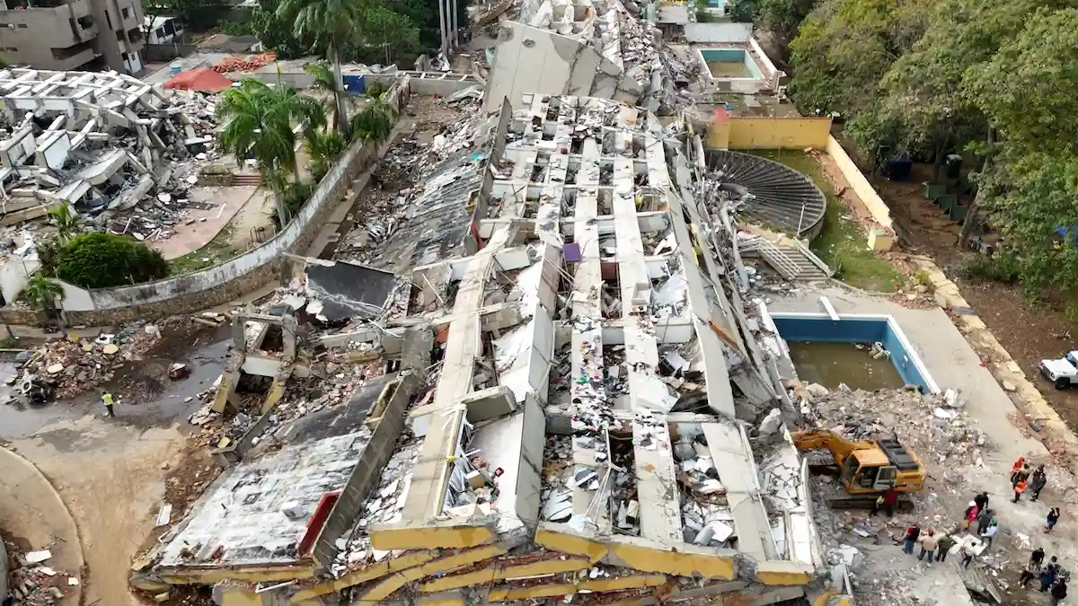 Vista aérea de un gran edificio de varios pisos completamente derrumbado y reducido a escombros. Se observa una excavadora amarilla trabajando en el lugar, personal de rescate y personas alrededor de las ruinas junto a una pileta vacía.