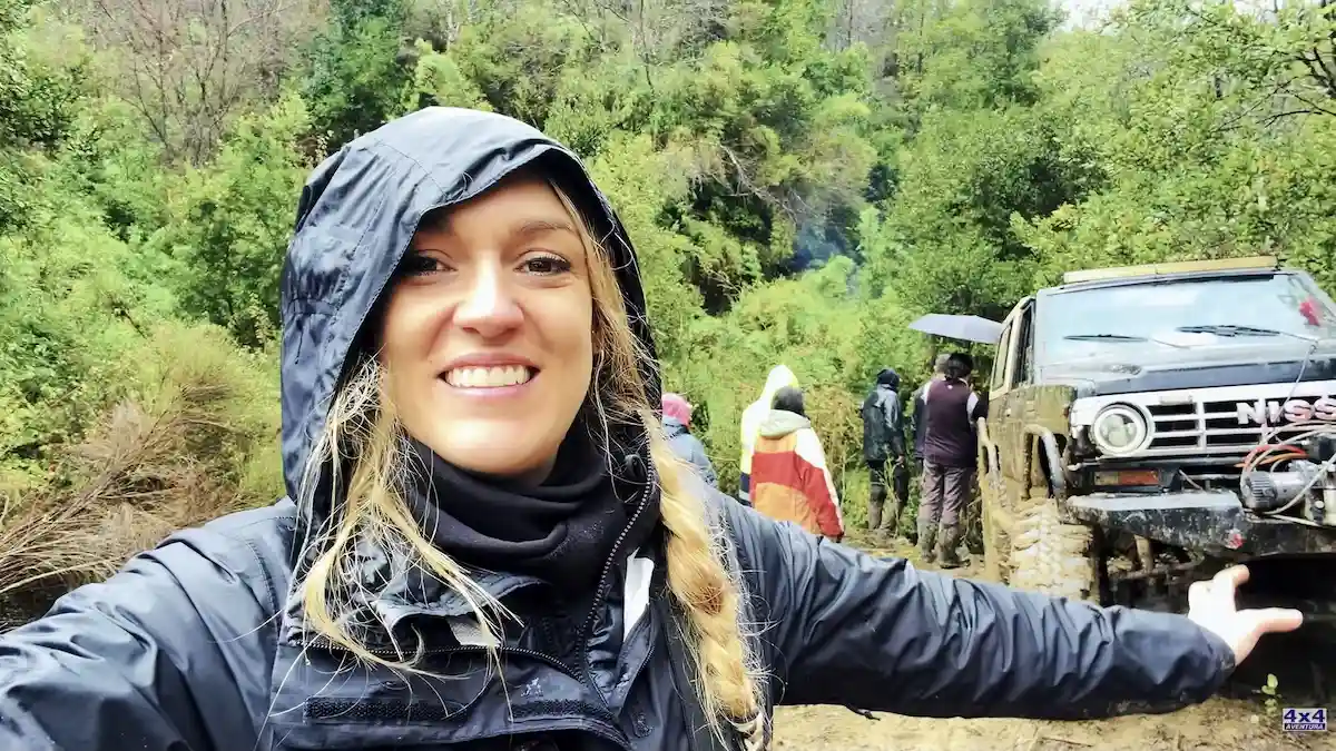 Texto alternativo: Mujer con capucha bajo la lluvia señalando una camioneta Nissan 4x4 en un camino de bosque.
