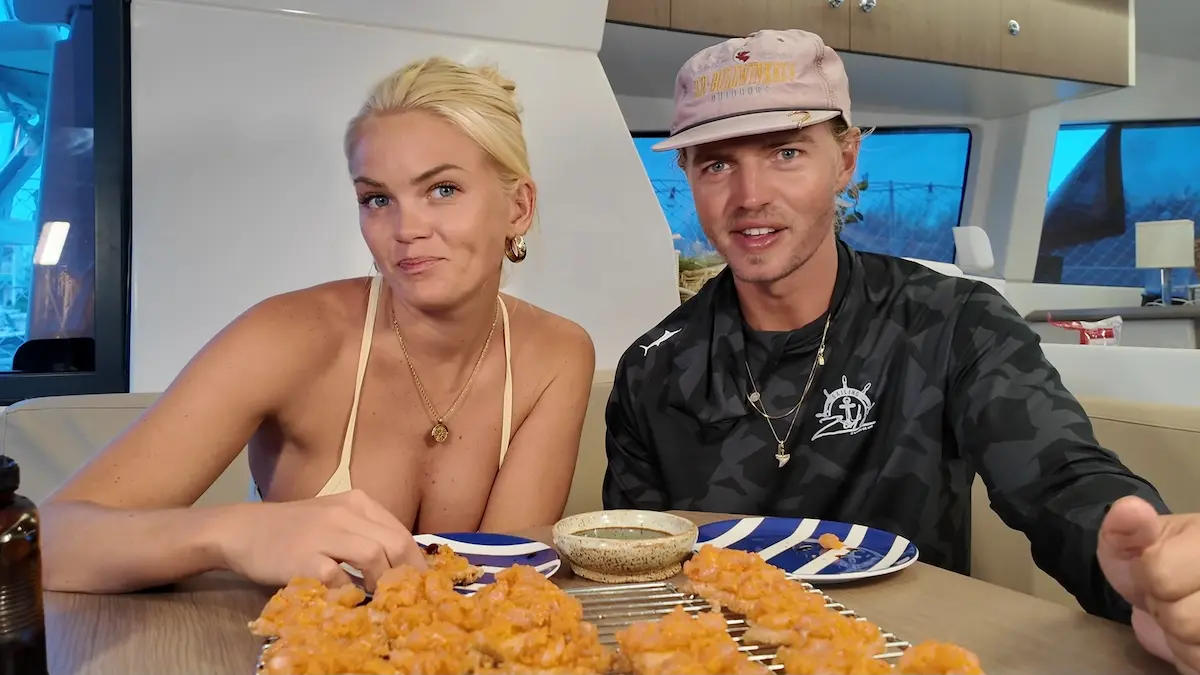 Pareja a bordo del catamarán lista para degustar bocados de pescado fresco condimentado sobre base crocante en la mesa del salón.