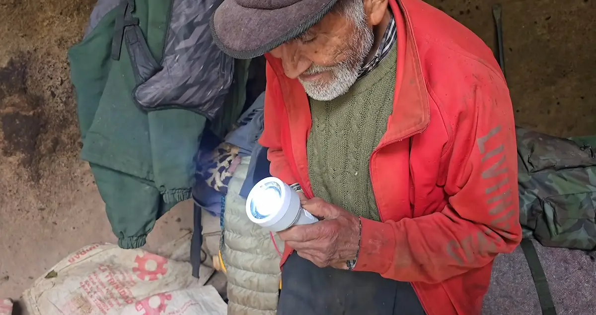 Primer plano de Don Pedro con barba y boina mirando una linterna solar blanca encendida que sostiene en sus manos dentro de su cueva