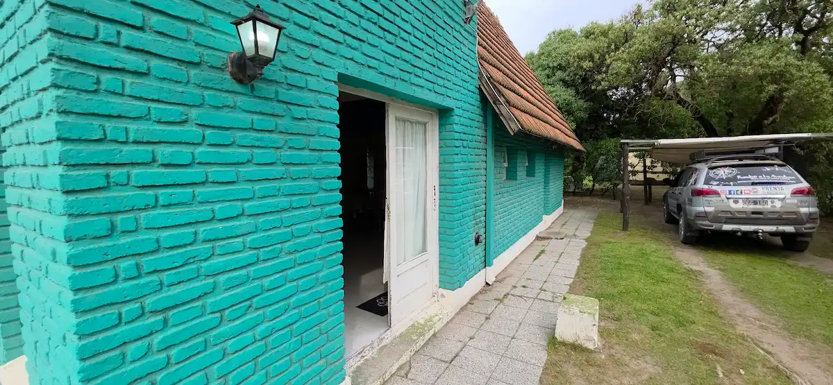 Vista exterior de una cabaña de ladrillo turquesa con farol encendido y puerta blanca abierta, mostrando la cochera techada con el SUV de prensa estacionado en el Complejo Camping Miguel Lillo de Necochea.[