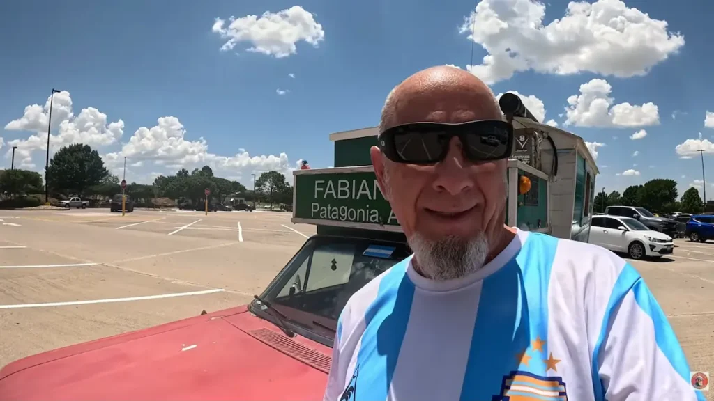 Un hombre de perfil aventurero, con anteojos de sol negros, barba candado canosa y vistiendo la camiseta de la Selección Argentina de fútbol, sonríe a la cámara en primer plano. Detrás de él se encuentra la parte delantera de una camioneta antigua de color rojo modificada con una estructura tipo casa rodante en la caja, la cual lleva un cartel verde donde se lee parcialmente "FABIAN Patagonia". El vehículo está estacionado en un amplio playón de estacionamiento al aire libre bajo un cielo azul brillante con nubes blancas dispersas.
