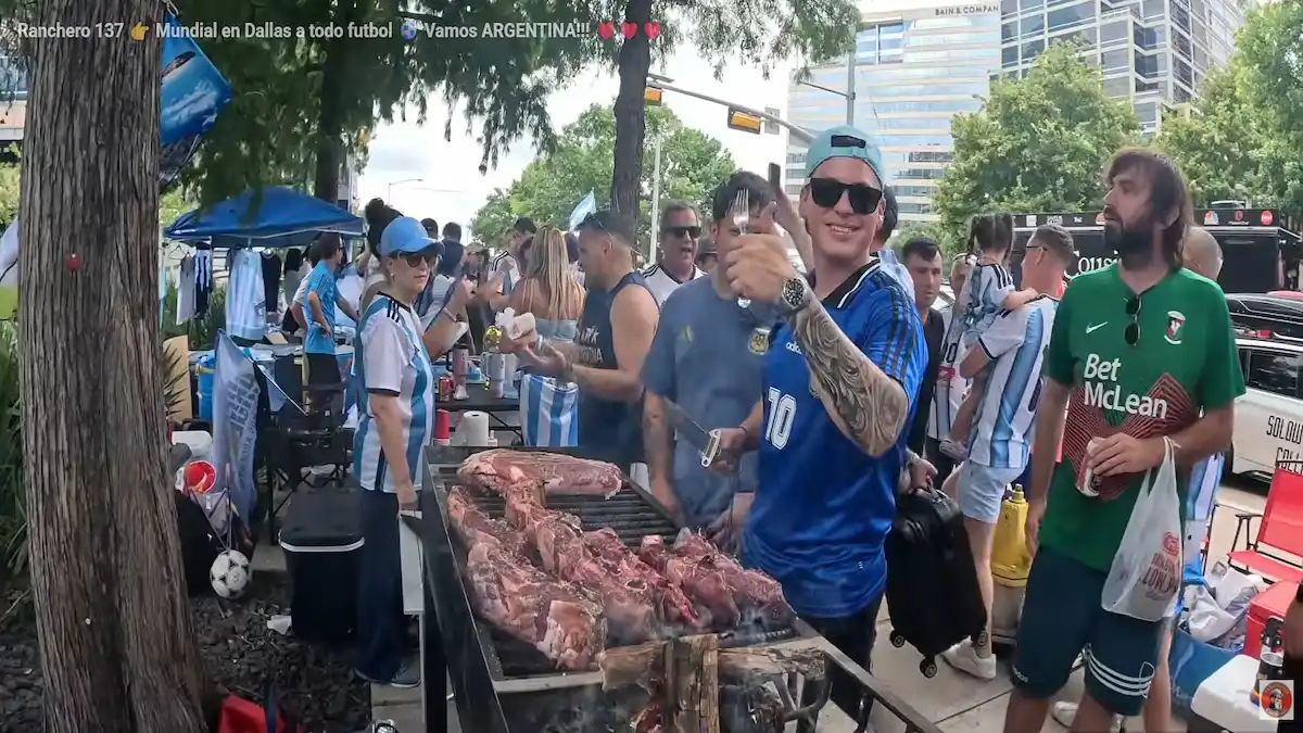 Una parrilla al aire libre repleta de costillares de carne vacuna y chorizos se cocina sobre las brasas durante el banderazo de los hinchas argentinos en Dallas, Texas. Alrededor, personas vestidas con camisetas de la Selección Argentina observan y comparten el tradicional asado en plena calle estadounidense.