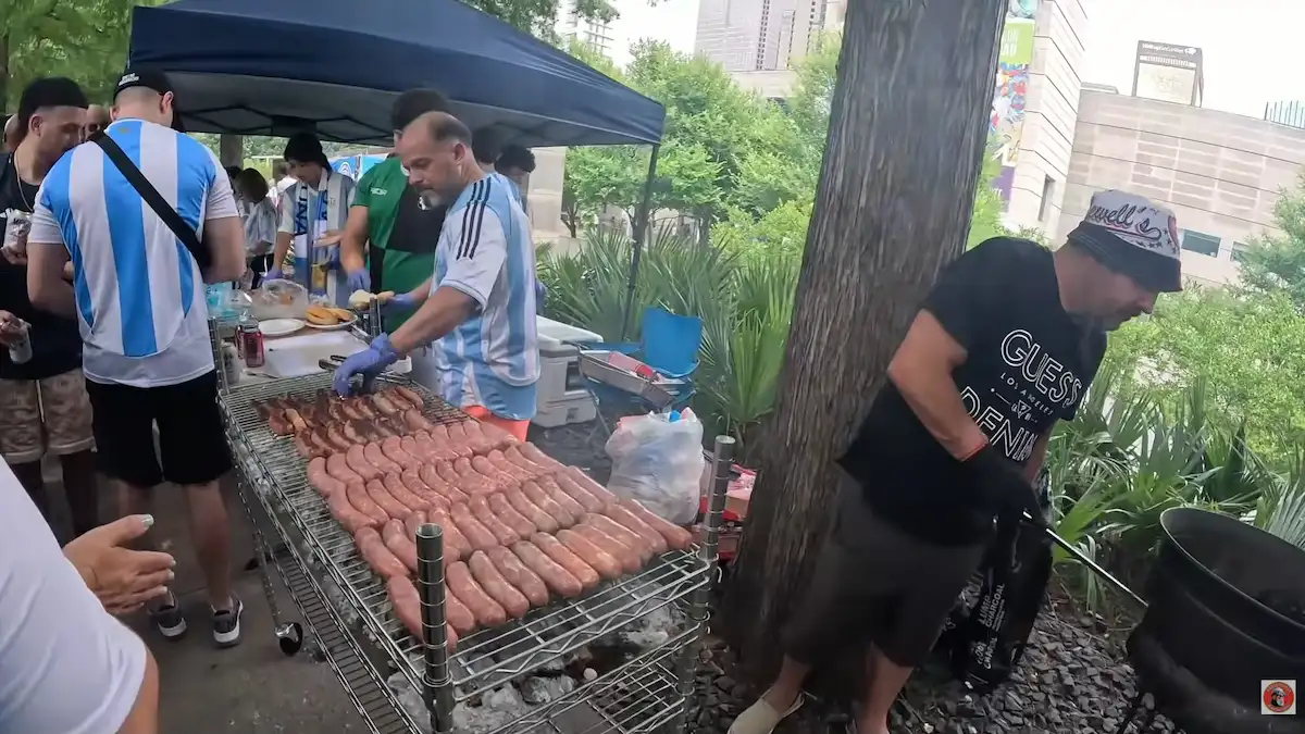 Parrilla llena de chorizos en Dallas Primer plano de una parrilla colmada con una gran cantidad de chorizos que se cocinan a las brasas sobre el fuego, desprendiendo humo al aire libre. La escena retrata el clásico almuerzo argentino adaptado en las calles de Dallas durante los festejos de la hinchada.
