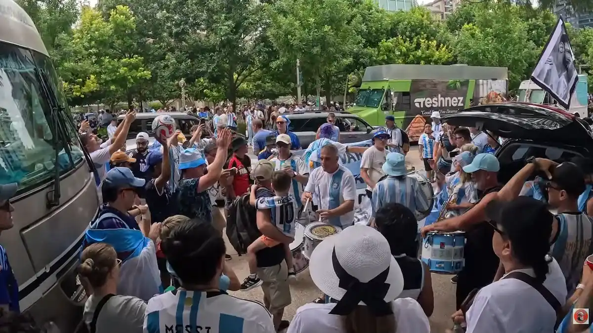Hinchada argentina con bombos en el Fan Fest de Dallas Una gran multitud de hinchas vestidos con la camiseta celeste y blanca de la Selección Argentina se agrupa al aire libre durante los festejos en Dallas. En el centro de la escena se destacan varios simpatizantes tocando bombos y redoblantes decorados con los colores nacionales, rodeados por banderas en alto y un ambiente de total euforia futbolera.