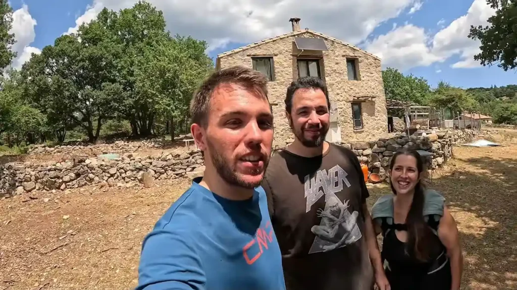 El creador de contenido Arnau junto a José y Sandra sonriendo frente a su casa sustentable de piedra y madera en medio de la montaña.