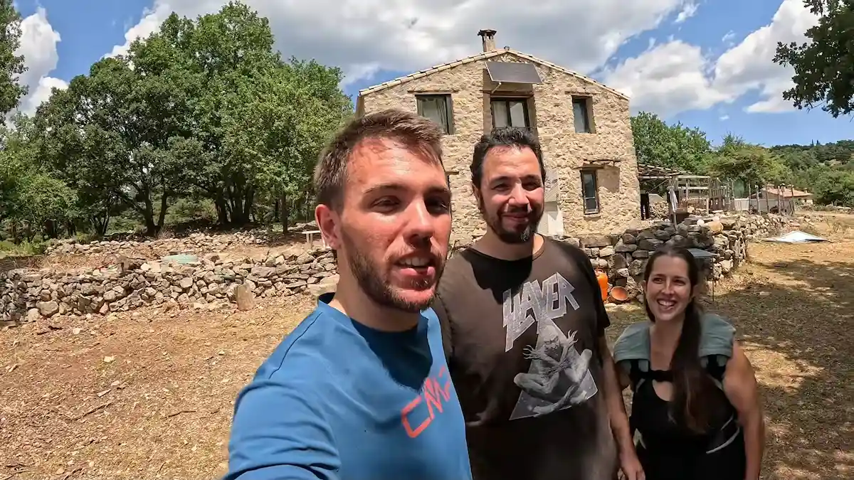 El creador de contenido Arnau junto a José y Sandra sonriendo frente a su casa sustentable de piedra y madera en medio de la montaña.