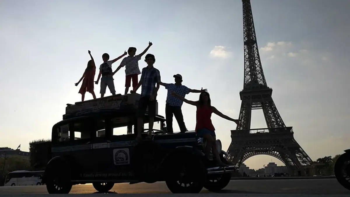 Una toma de silhouette en color invertido de la familia Zapp al completo (seis miembros: cuatro niños y dos padres) posando sobre su auto clásico Graham-Paige 1928, con el auto, las figuras y la Torre Eiffel en primer plano en tonos claros y brillantes, contra un cielo oscuro, profundo y de aspecto nocturno. Se aprecian detalles invertidos de la vestimenta y la estructura de la torre.
