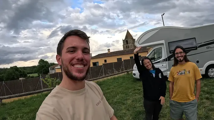 : El creador digital Arnau Serrado en primer plano sonriendo a la cámara y señalando con el dedo hacia arriba, mientras de fondo la pareja viajera de Furgo en Ruta, Marta y Marià, sonríen junto a su motorhome de Wow Campers en un entorno natural bajo un cielo nublado.