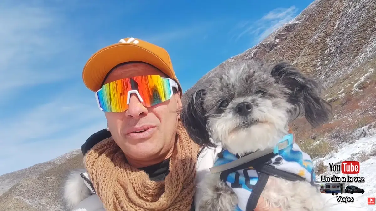 Javier, embajador de la Red Rumbo, sonríe a la cámara mientras sostiene a su perro en brazos frente a las montañas nevadas de Vallecitos en Mendoza.