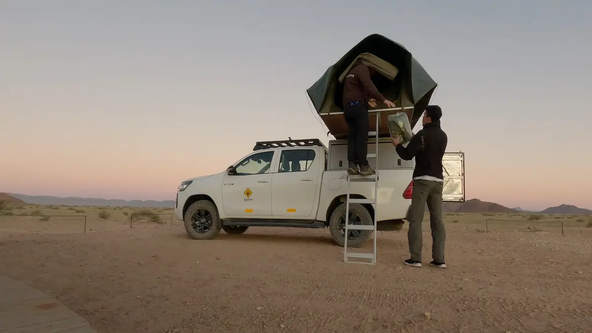 Dos hombres armando una tienda de campaña sobre el techo de una camioneta pickup en el desierto de Namibia al atardecer.