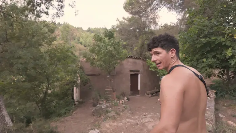 Mark de espaldas señalando la antigua barraca de piedra restaurada rodeada de vegetación en una finca de Cataluña.