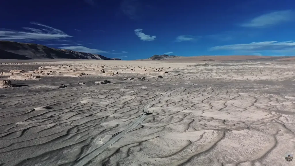 Vista aérea de una camioneta 4x4 circulando por una huella solitaria en medio de un enorme desierto de formaciones rocosas y dunas que asemejan un paisaje de Marte, bajo un cielo azul en la Puna de Catamarca.