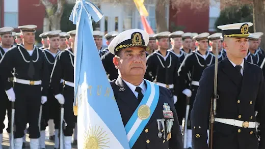 Un oficial de la Prefectura Naval Argentina en uniforme de gala sostiene la bandera nacional durante un acto oficial, con una formación de marineros alineados al fondo.