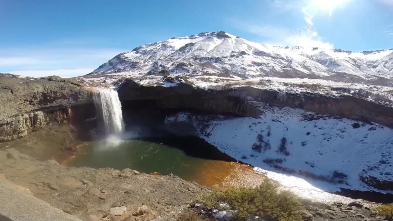 Vista panorámica de la Cascada del Agrio en Caviahue, Neuquén, con agua cayendo a una laguna verde esmeralda y montañas nevadas al fondo bajo un cielo azul despejado y sol brillante.
