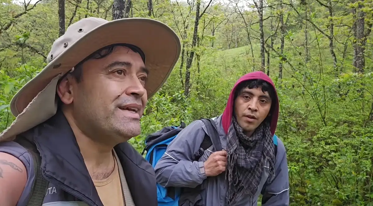 Sergio Caballero con sombrero y su compañero Gabriel con mochila caminando por el monte durante la travesía hacia la cueva de Don Pedro