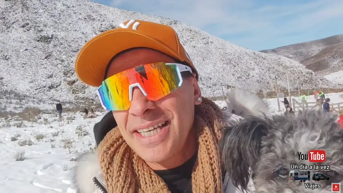 Un hombre con campera roja sonríe mientras sostiene en brazos a su pequeño perro, ambos posando frente a un majestuoso paisaje de la Cordillera de los Andes cubierta de nieve en Vallecitos, Mendoza.