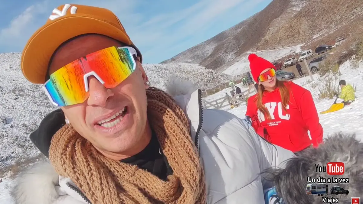 Primer plano de Javier con gorra, gafas de sol espejadas y bufanda, junto a Fátima vestida de rojo y su perro, en un parque de nieve en Vallecitos,